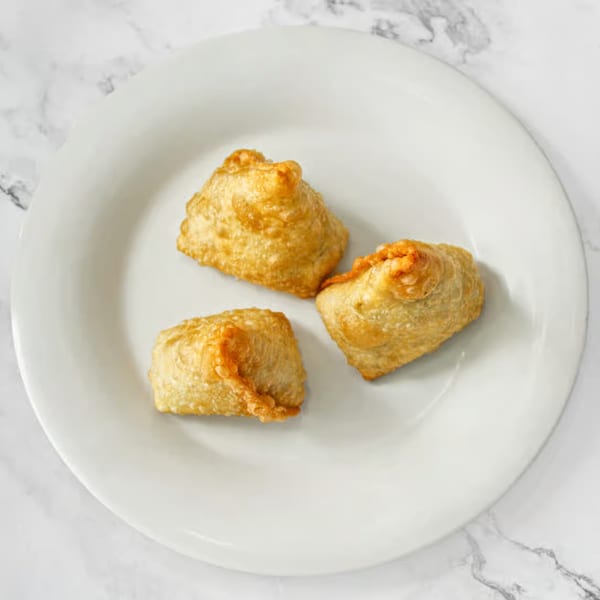 Three golden-brown, fried Indian samosas are arranged on a white plate against a marble background.