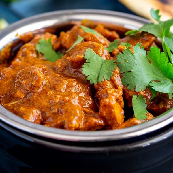 Close-up of a bowl of Indian curry, rich orange-brown with chicken pieces and fresh green cilantro.