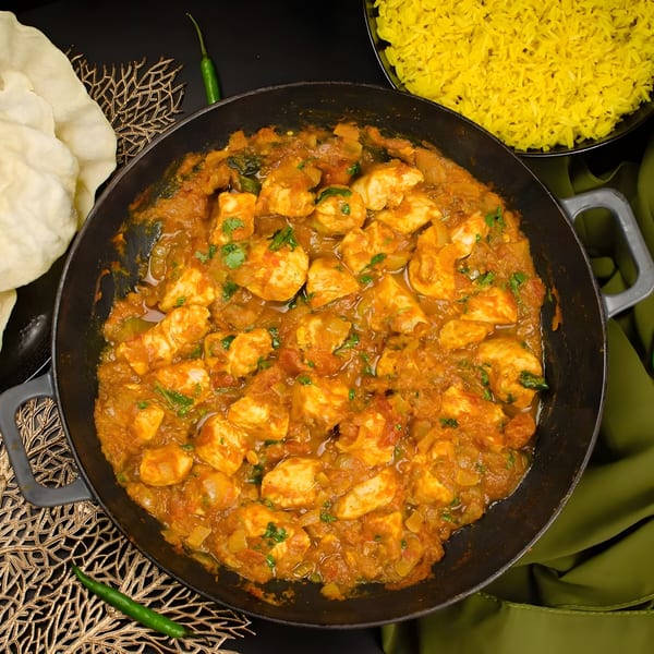 Indian chicken curry in a black pan with yellow rice, papadum, and green chilies.