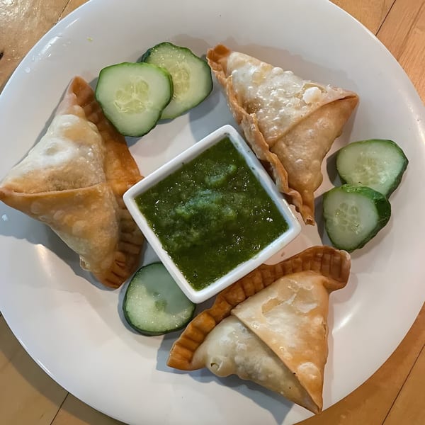 Four golden-brown samosas with a side of green chutney and cucumber slices on a white plate.